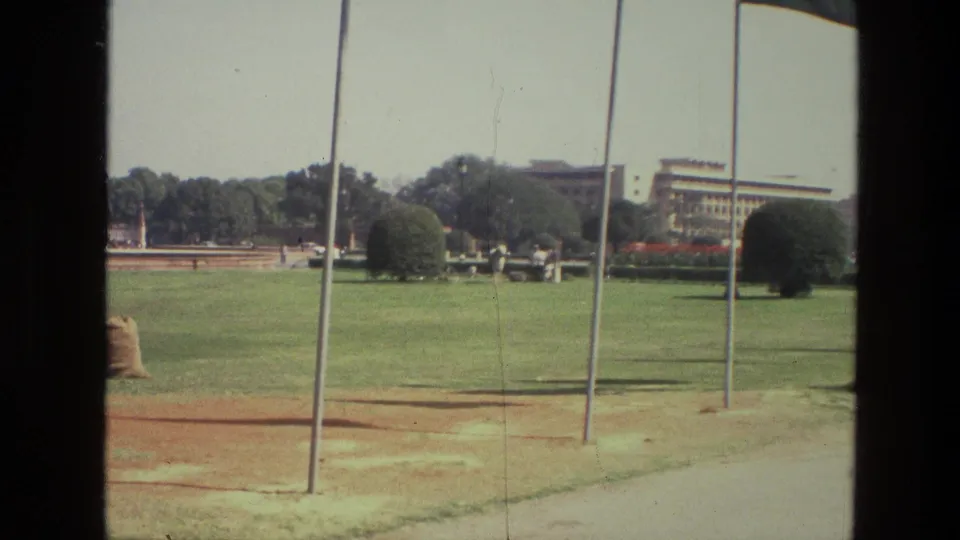 1974 India 8mm frame showing flagpoles, a paved path, open lawn, trimmed shrubs and a large building in the distance.