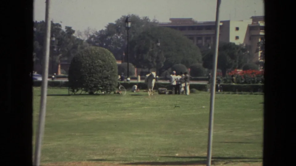 1974 India 8mm frame showing several people on a manicured lawn near a white horse, rounded shrubs and a distant building.