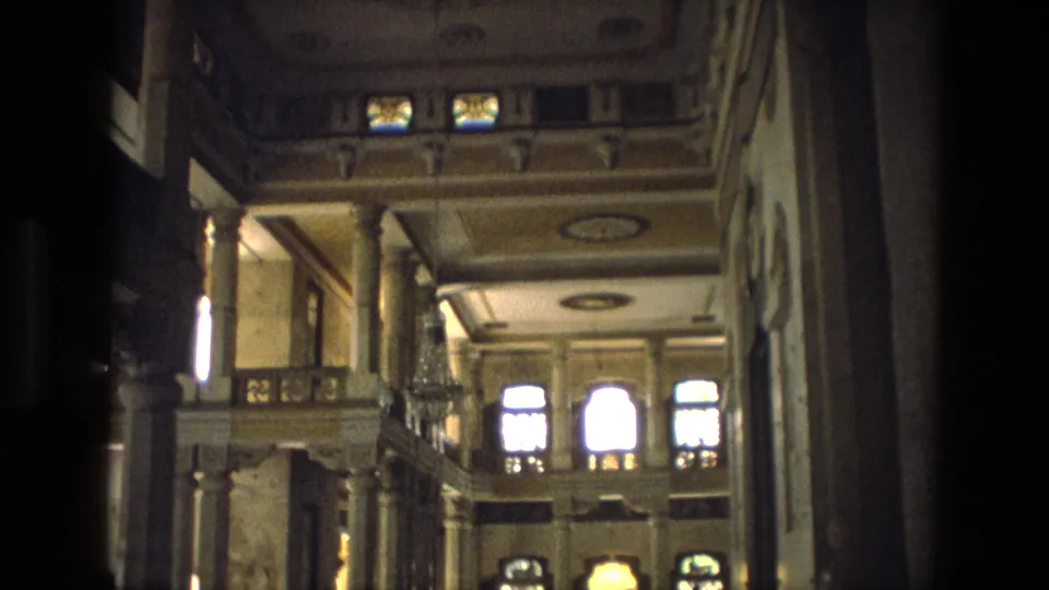1974 India home-movie frame showing a grand palace interior with columns, upper balcony railings, arched windows, and soft interior light.