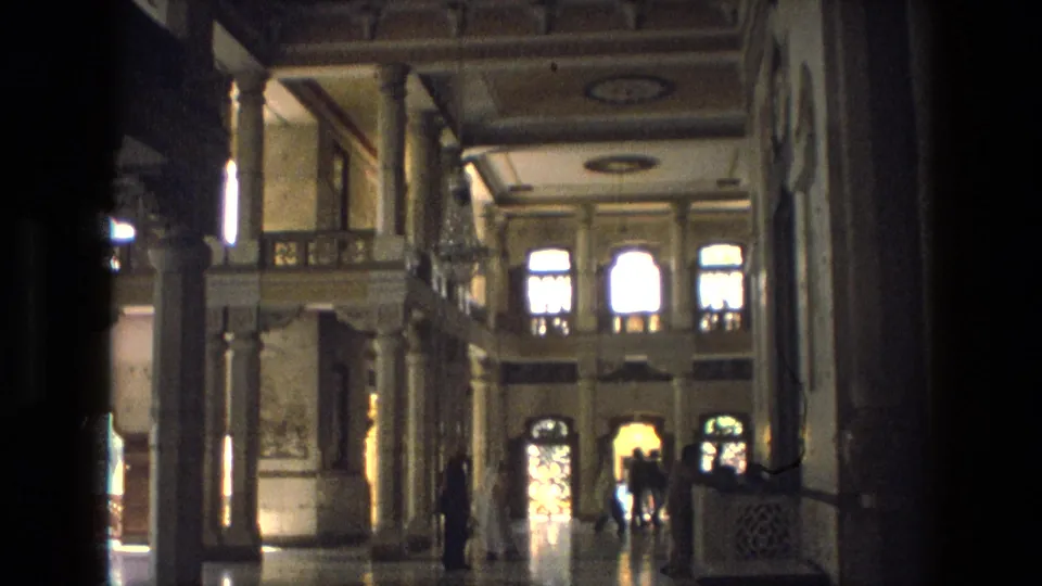 1974 India home-movie view inside an ornate palace hall, showing columns, reflective flooring, arched windows, and distant visitors.