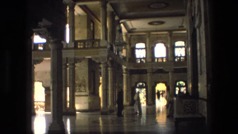 1974 India frame showing a wide palace interior with columns, patterned ceiling panels, arched windows, and distant visitors.