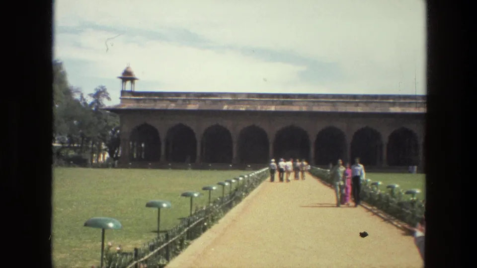 1974 India home-movie frame showing visitors on a concrete walkway approaching a broad arched building across formal lawns.
