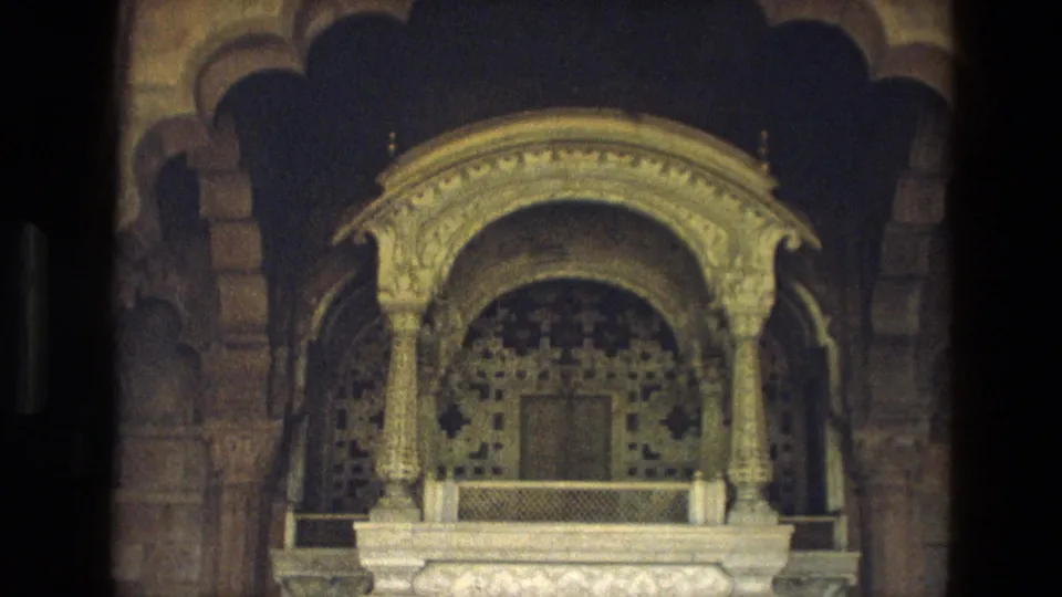 1974 India home-movie frame of an ornate interior niche with carved columns, patterned wall detail, and a low protective barrier.