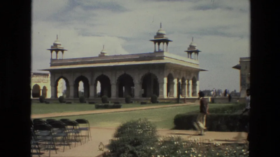 1974 India home-movie view of a pavilion across manicured grounds, with folding chairs in front and visitors near the walkway.