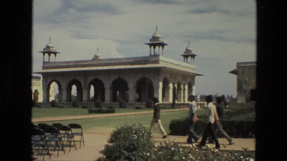 1974 India exterior frame with several visitors crossing a garden path before a domed, arched pavilion and trimmed hedges.