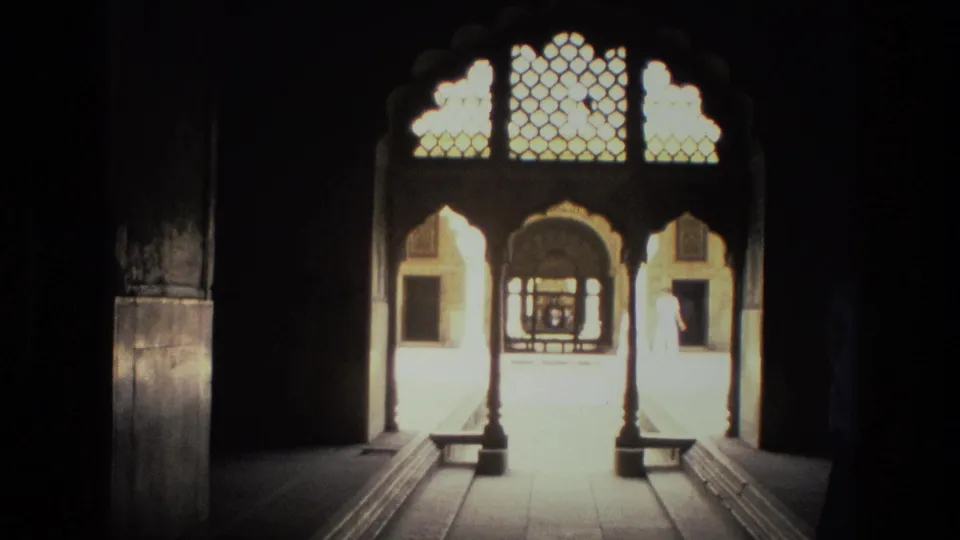 From a shadowed interior, the camera looks toward a bright arched gateway with carved lattice panels in India.