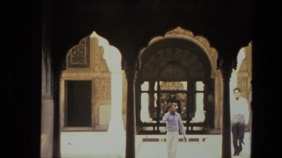 1974 India 8mm frame with a man walking across a sunlit courtyard, viewed through dark ornate arches and latticework.