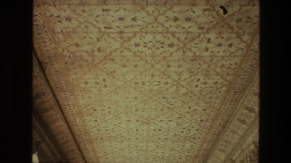 1974 India 8mm frame showing a palace ceiling viewed upward, with patterned ornament, angled borders, and a small bright light patch.