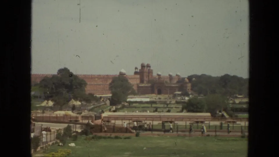 1974 8mm home-movie view in India showing a distant Mughal-era red sandstone fortress beyond gardens, paths, trees, and small visitor figures.