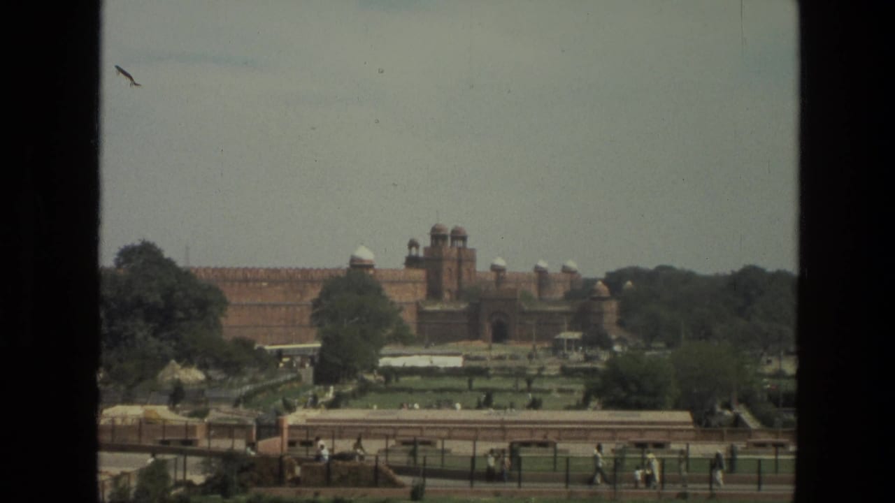 1974 India footage frames a broad red sandstone fortress with domed towers behind green gardens, walkways, trees, and scattered visitors.