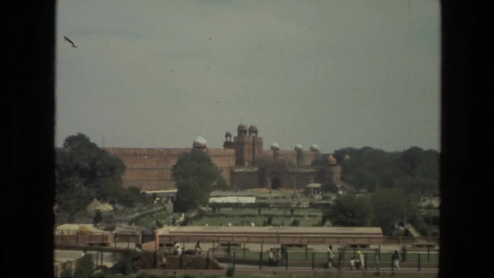 1974 India footage frames a broad red sandstone fortress with domed towers behind green gardens, walkways, trees, and scattered visitors.