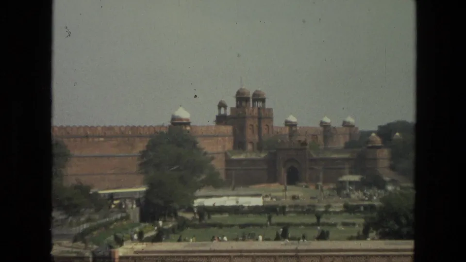 A closer 1974 8mm view in India shows red sandstone walls, domed towers, an arched entrance, gardens, trees, and small figures in the grounds.