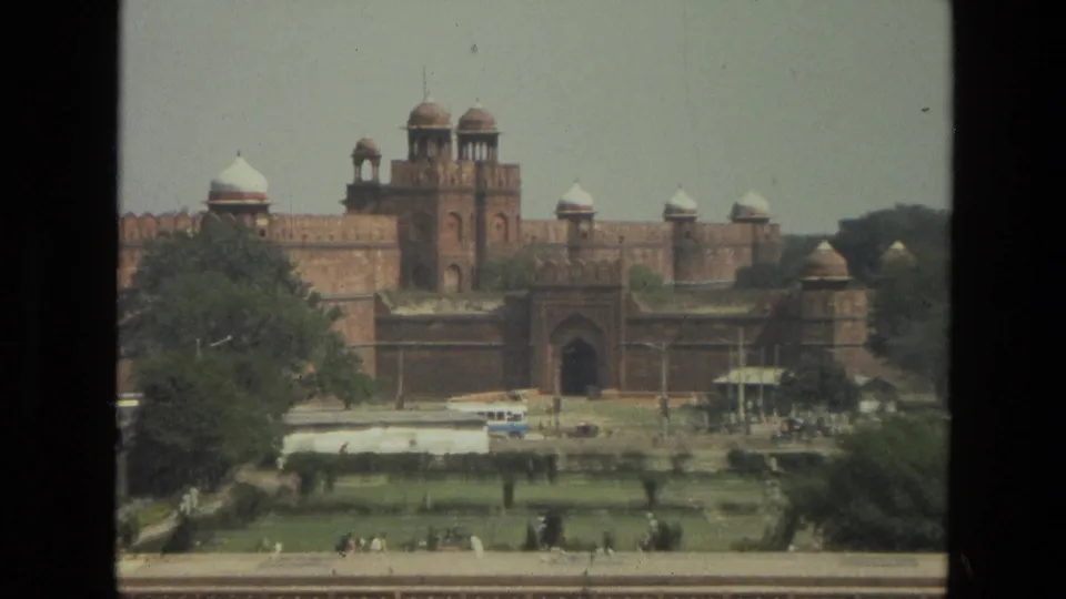 1974 India home-movie frame centers the red sandstone fortress gateway and domed towers above green gardens, trees, paths, and distant visitors.