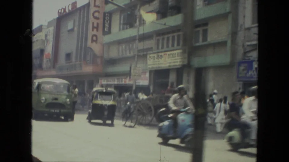 1974 Indian city street with mixed traffic, scooters, an auto-rickshaw, pedestrians, and Hindi storefront signage.