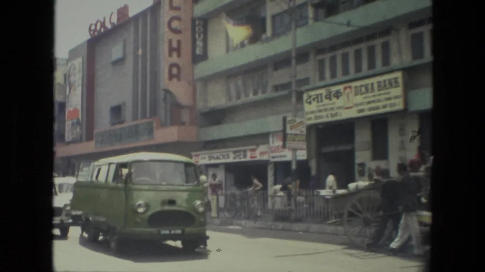 1974 Indian street scene with a green vehicle, shopfronts, Hindi signage, pedestrians, and a cycle rickshaw at the curb.
