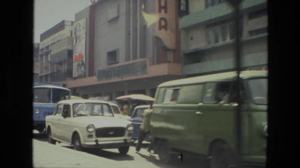 1974 Indian urban road with a white car, green van, bus, pedestrians, and commercial building fronts in soft 8mm color.