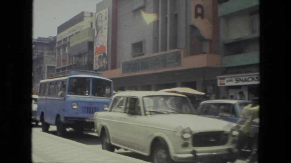 1974 India street scene with a blue bus, pale vintage car, traffic, and storefronts seen from a handheld 8mm home movie.