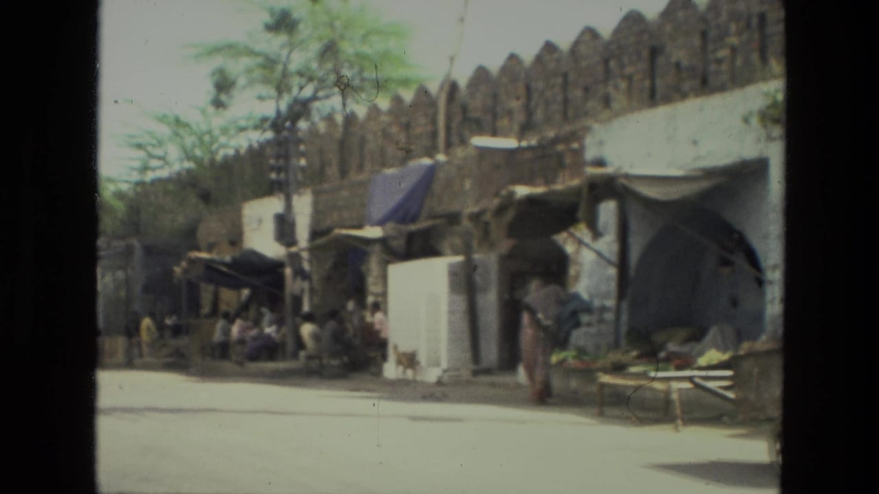 Roadside stalls and pedestrians appear beside a stone wall in 1974 India, filmed with soft handheld 8mm texture.