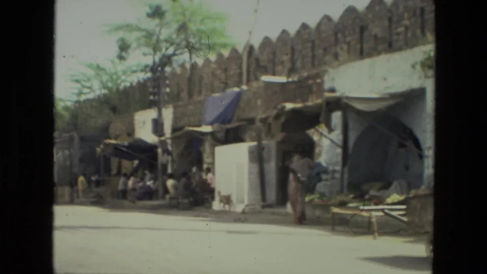 Roadside stalls and pedestrians appear beside a stone wall in 1974 India, filmed with soft handheld 8mm texture.
