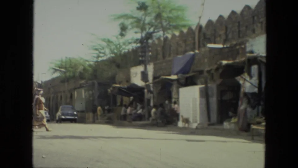 A woman crosses a sunlit street near roadside stalls, a stone wall, and parked vintage cars in 1974 India.