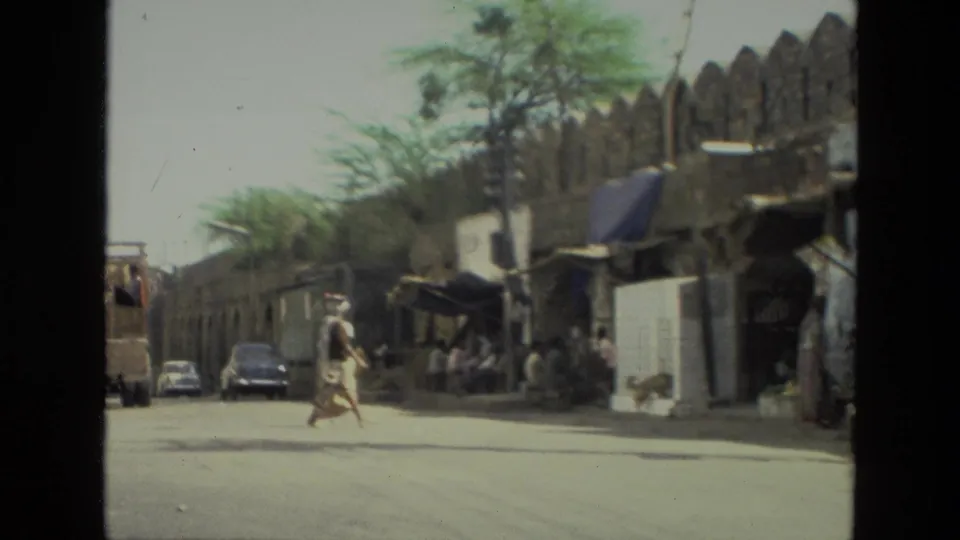 Handheld 8mm frame of a woman crossing a road in 1974 India, with shop awnings, pedestrians, and vintage cars nearby.