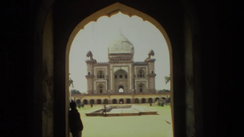 1974 India 8mm frame of a grand domed monument and courtyard viewed through a shaded arched gateway, with a visitor silhouette for scale.