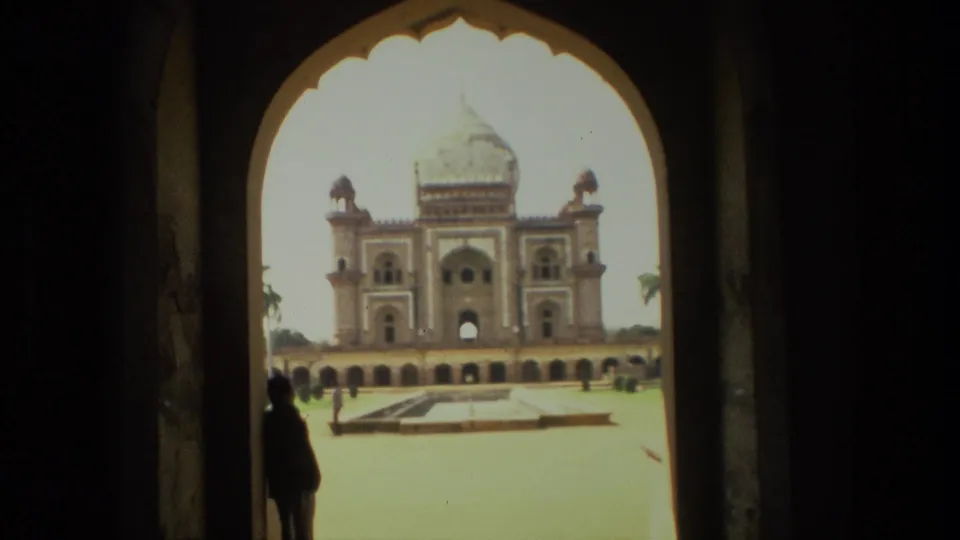 Visitor-shot 1974 India view of a Mughal-style domed structure beyond a courtyard, framed by a dark arch and a standing silhouette.