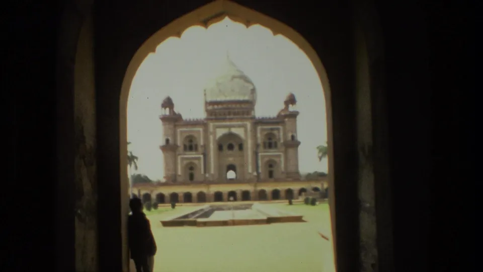 Faded 8mm frame from India showing a domed historic structure across a sunlit courtyard, partially framed by a dark entrance arch.