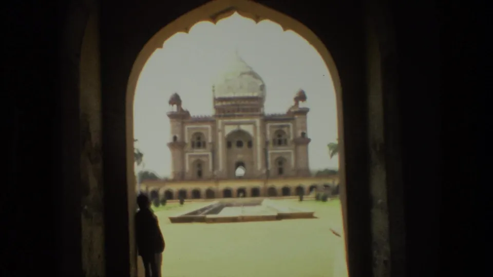 1974 India home-movie still of a monumental domed facade beyond a bright courtyard, viewed from inside a shaded arched gateway.