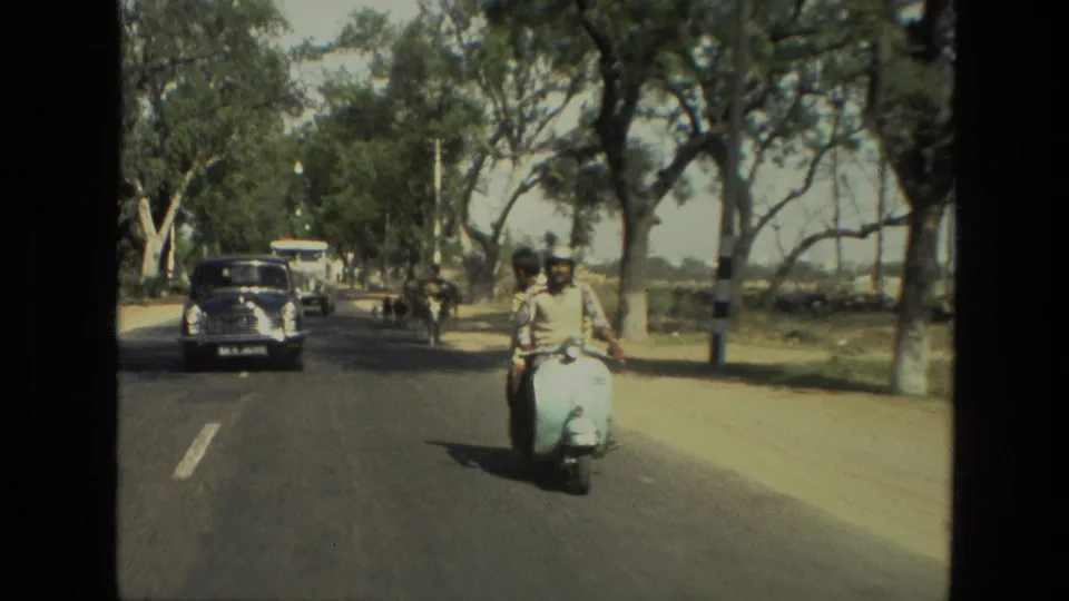 1974 India traffic frame with a scooter in the foreground, older vehicles behind, and goats farther along the road.