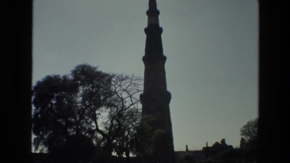 1974 Delhi home-movie frame showing Qutub Minar silhouetted above trees against a bright sky.