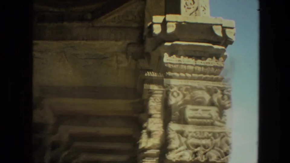 1974 India home-movie frame with a sunlit carved stone column, decorative reliefs, and ruin masonry against blue sky.