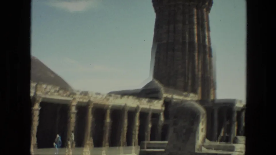 1974 India 8mm frame of a temple courtyard with columns, a tall stone structure, and distant visitors walking through the site.
