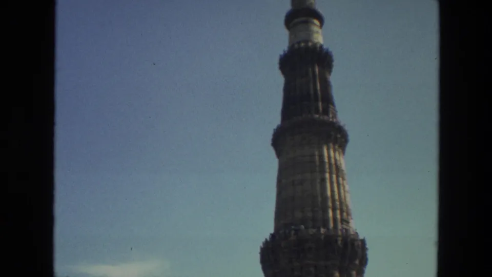 1974 Delhi home-movie frame showing the upper Qutub Minar tower and balcony details beside a dark window-frame edge.