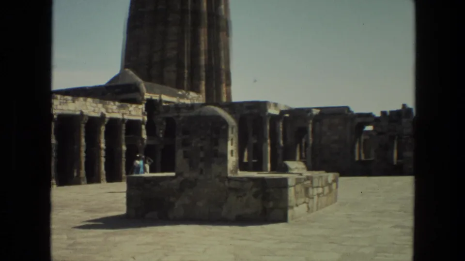 1974 8mm view of Qutub Minar grounds in Delhi, showing stone paving, ruins, colonnades, and small visitors.