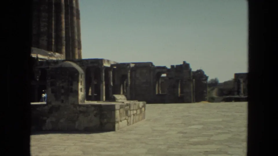 1974 Delhi home-movie frame of Qutub Minar grounds, with open stone paving, ruins, tower base, and a small visitor.