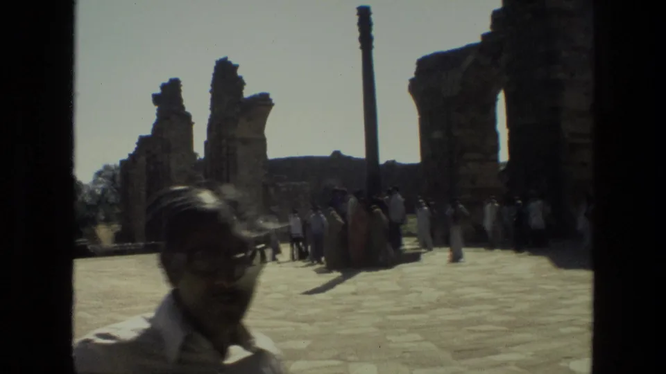 1974 India 8mm frame with a visitor close to camera and a group gathered near tall stone ruin remains.