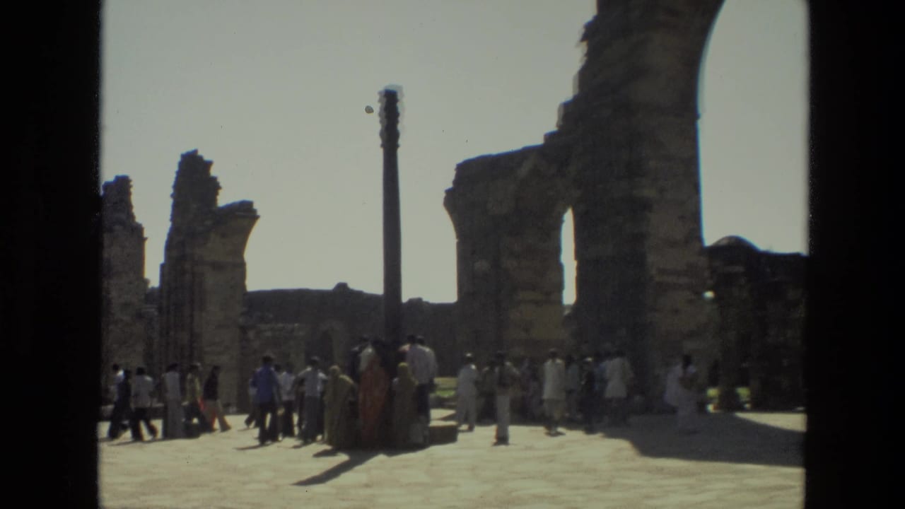 Tourists walk and gather near weathered arches and a pillar in 1974 home-movie footage from Delhi's Qutub Minar complex.
