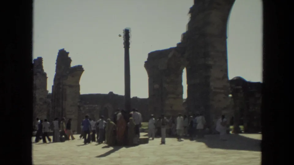 Tourists walk and gather near weathered arches and a pillar in 1974 home-movie footage from Delhi's Qutub Minar complex.