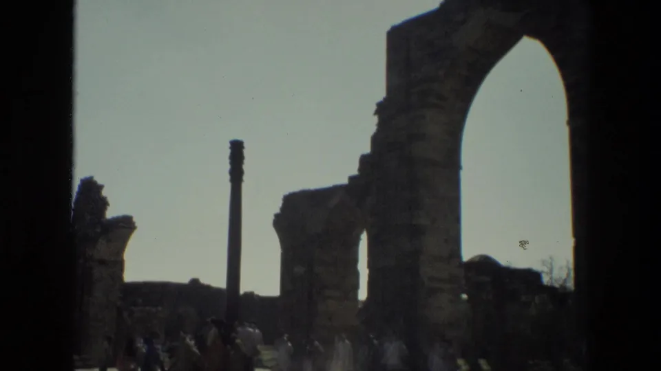 A handheld 8mm frame shows stone arches, a tall pillar, and distant visitors at the Qutub Minar ruins in India.