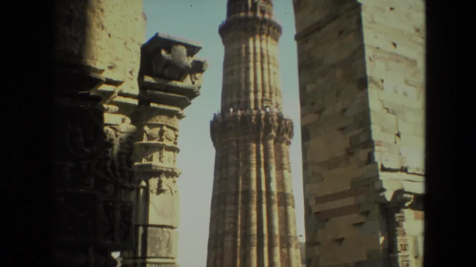 The minaret is filmed from inside the Qutub complex, with stone columns and inscriptions partly visible around it.