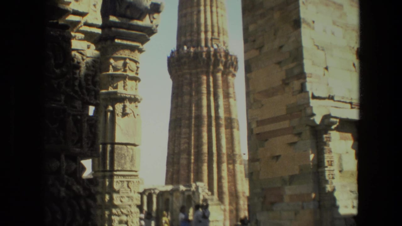 1974 Delhi travel footage frames Qutub Minar through dark stone columns, with visitors standing near the historic structure.