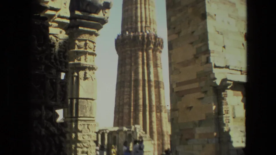 1974 Delhi travel footage frames Qutub Minar through dark stone columns, with visitors standing near the historic structure.
