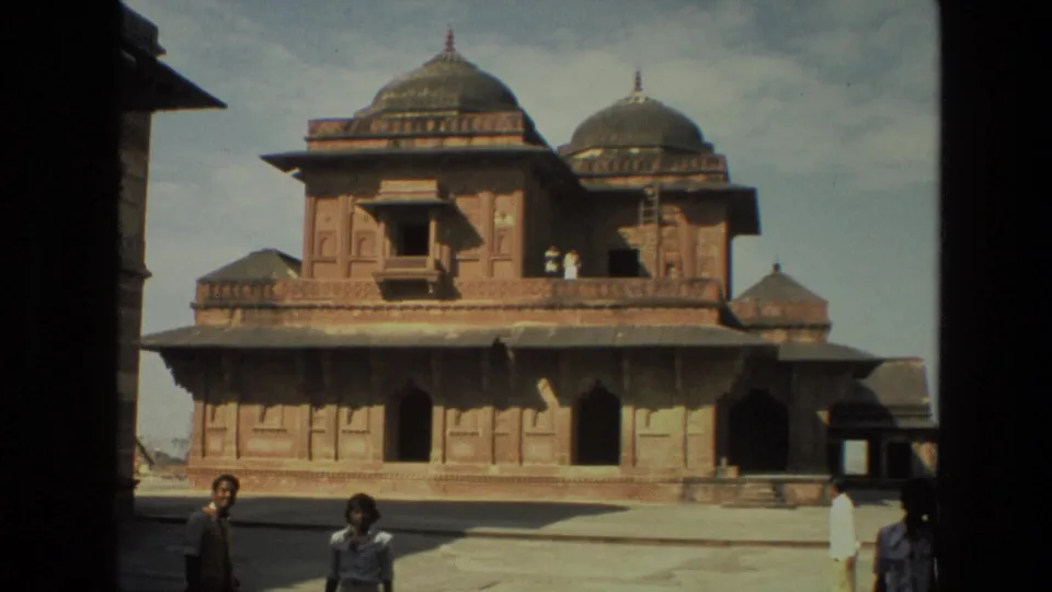 A red sandstone domed building in the Delhi historic complex is shown across a courtyard, with several visitors walking nearby.