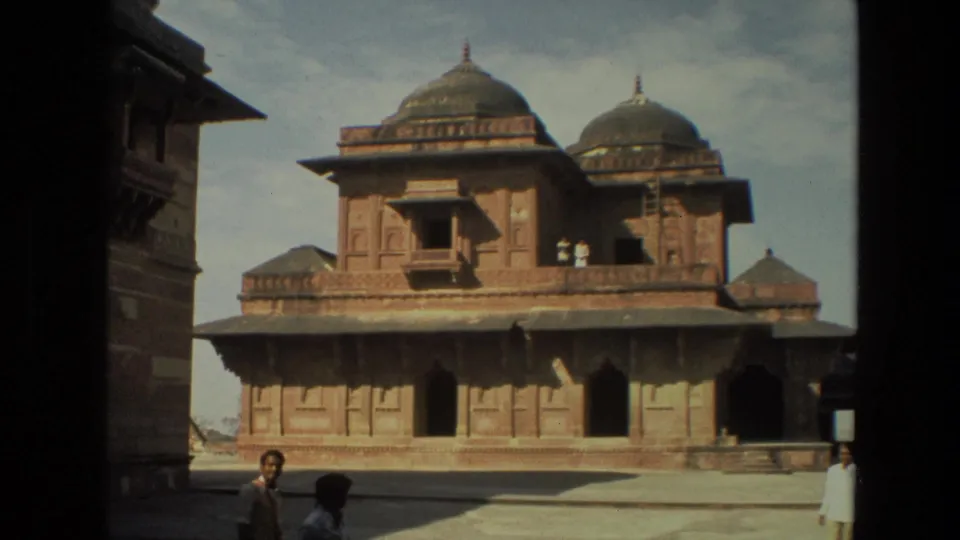 The 8mm travel view holds on a domed red sandstone building in Delhi, with tourists partly visible around the courtyard.