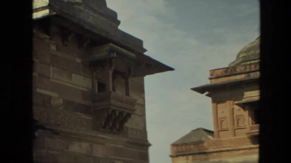 1974 India home-movie frame with partial Mughal-style domed architecture, red sandstone carving, shaded framing, and bright sky.