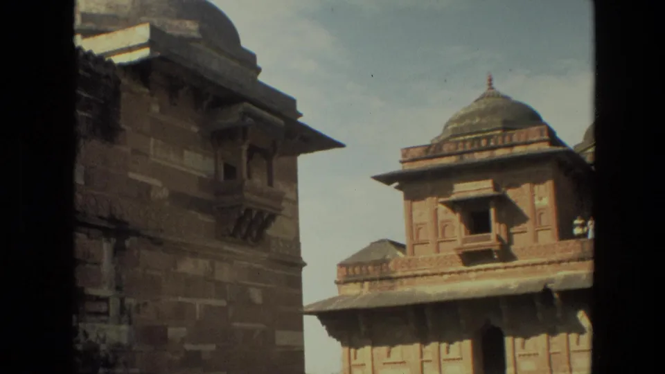 1974 India home-movie frame showing carved red stone architecture and domed rooflines from a moving visitor viewpoint.