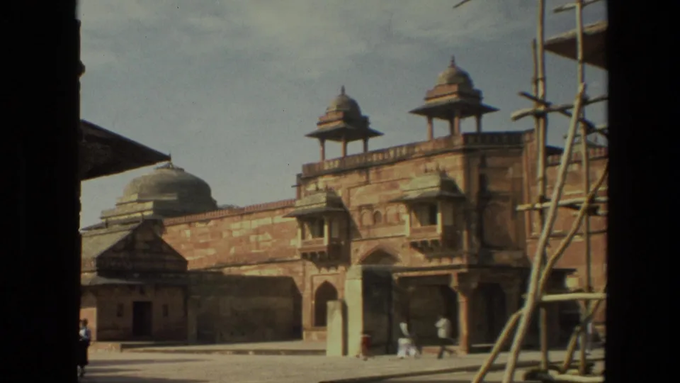 1974 8mm India travel frame showing a historic palace courtyard with sandstone walls, domed pavilions, scaffolding, and distant visitors.