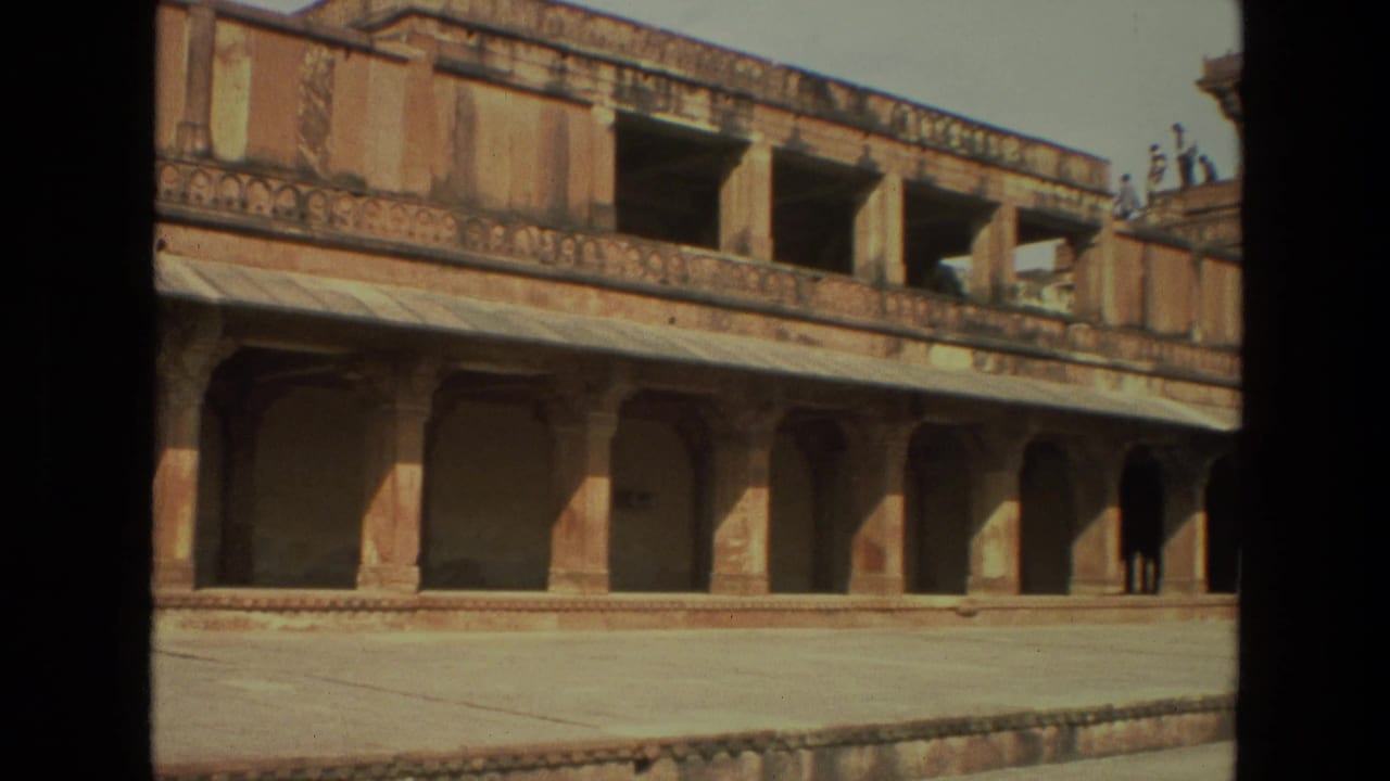 1974 India 8mm frame of a palace facade with arched colonnades, upper balcony openings, carved railing, and sunlit courtyard paving.