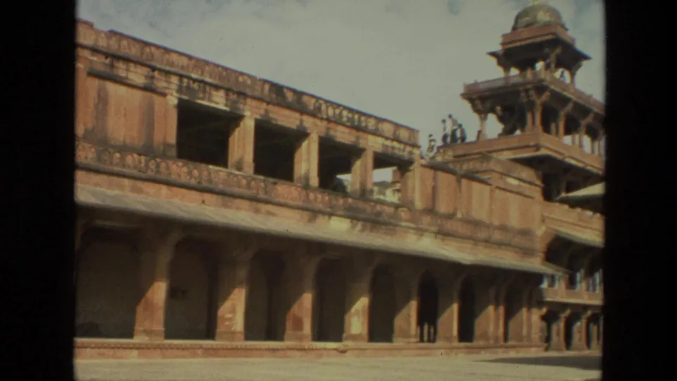 1974 8mm India frame showing a sandstone palace facade, arched walkway, projecting pavilion tower, and small visitors near the courtyard edge.